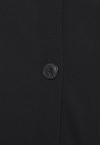 Musta neuletakki, jossa on teksturoitu neulos ja etuosassa yksi musta nappi, yksinkertainen muotoilu ja käytännöllisyys.