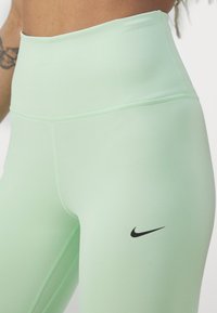 Jasnozielone legginsy z wysokim stanem, wykonane z gładkiego materiału, z czarnym logo Nike na dolnej części i tonalnymi detalami szycia.
