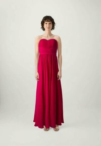 Garais, bezplecu vakara kleita spilgti rozā audumā. Ar pieguļošu korsešu, kas satur krokas, un plūstošu svārku. Valkāta ar gaišas krāsas sandālēm.