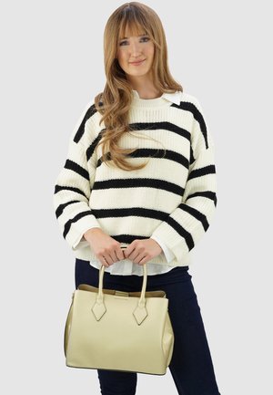 Junge Frau mit langen Haaren, die einen weiß-schwarz gestreiften Pullover und dunkle Hosen trägt und eine beigefarbene Handtasche mit beiden Händen hält.