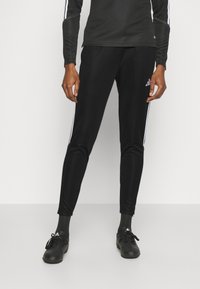 Čierne športové nohavice s bielymi bočnými pruhmi, úzky strih, vyrobené z ľahkého materiálu. Kombinované s čiernymi topánkami a ponožkami.