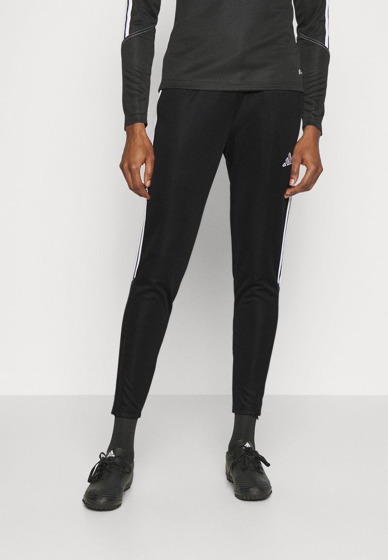 Čierne športové nohavice s bielymi bočnými pruhmi, úzky strih, vyrobené z ľahkého materiálu. Kombinované s čiernymi topánkami a ponožkami.