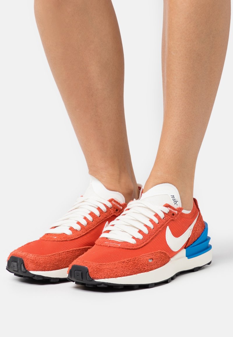 Gros plan sur des pieds portant des baskets Nike orange et blanches avec des accents bleus au talon et des lacets blancs sur fond blanc.