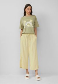 Oliwkowa koszulka z białym wzorem, zestawiona z jasnokremowymi spodniami o szerokich nogawkach. Modelka nosi białe sandały na platformie.