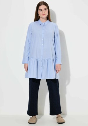 Abito camicia azzurro chiaro a maniche lunghe con chiusura a bottoni e fondo arricciato, abbinato a pantaloni scuri a fantasia e scarpe beige.
