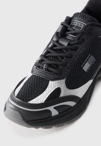 Black athletic shoe featuring a mesh upper with glossy accents, a thick sole, and braided laces. Silver panels and logo detail included.