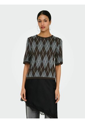 Donna con capelli scuri che indossa orecchini a cerchio, maglione a maniche corte con motivo a rombi e gonna nera asimmetrica con bordo in pizzo sopra jeans.