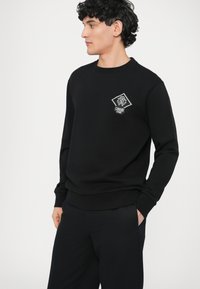 Czarna bluza z okrągłym dekoltem, ściągaczami na mankietach i na dole. Na piersi znajduje się biała naszywka w kształcie rombu. Gładka faktura materiału.