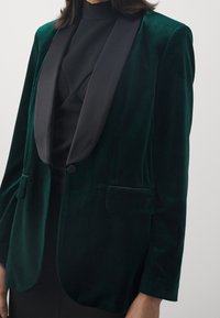 Sammet grön blazer med svarta satinlapeller, enkelknappsstängning, två framlommer och en slät texturfinish.