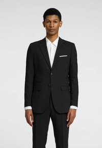 Chaqueta de traje negra con dos botones, solapas de muesca y bolsillos delanteros. Llevada sobre una camisa blanca de botones con cuello puntiagudo.