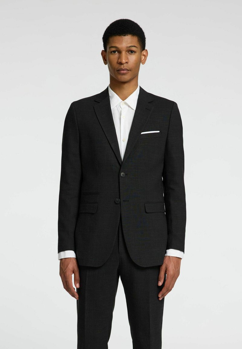 Chaqueta de traje negra con dos botones, solapas de muesca y bolsillos delanteros. Llevada sobre una camisa blanca de botones con cuello puntiagudo.