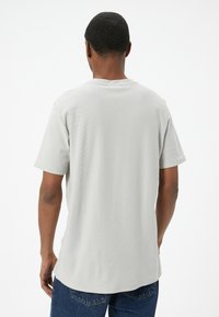 Camiseta de manga corta color gris claro con un cuello redondo y una tela texturizada. La parte de atrás presenta un dobladillo ligeramente más largo con un corte recto.