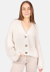 Strukturierte Creme-Strickjacke mit übergroßen Ärmeln, V-Ausschnitt und drei metallischen Knöpfen vorne, getragen über einem hellen Top und kombiniert mit beigen Hosen.