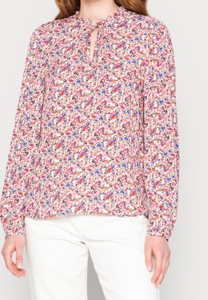 Vrouw draagt een bloes met lange mouwen met bloemenprint in roze, rood, blauw en beige, gecombineerd met een witte broek.