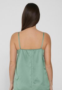 Camisole en satin de couleur vert doux, avec des bretelles ajustables, un dos froncé et une texture douce avec une coupe légèrement loose.
