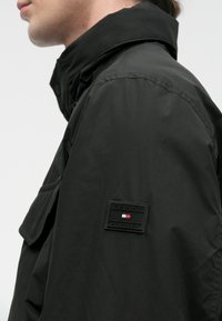 Mustapadded-takki korkealla kauluksella, jossa on suorakaiteen muotoinen logo-patch mustana, punaisena ja valkoisena. Sujuva pinta ja käytännöllinen muotoilu.