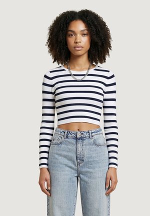 Giovane donna con capelli ricci che indossa una maglietta a maniche lunghe corta a righe bianche e blu navy e jeans a vita alta di colore azzurro chiaro.