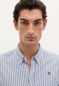 Blau-weiß gestreifte Button-Down-Hemd, Baumwollstoff, klassischer Kragen, kleines Totenkopf- und Kreuzknochels-Logo auf der linken Brust.