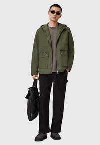 Veste à capuche vert olive avec deux poches avant, t-shirt gris en dessous, pantalon en velours côtelé noir et un sac noir. Porté avec des chaussures sombres et des lunettes de soleil.