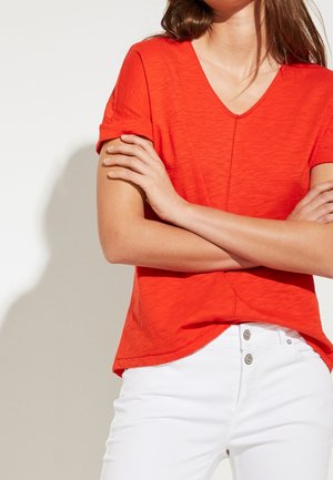 Frau trägt ein hellrotes, kurzärmeliges V-Ausschnitt-Oberteil mit aufgerollten Manschetten, kombiniert mit hoch taillierten weißen Hosen, Arme verschränkt.