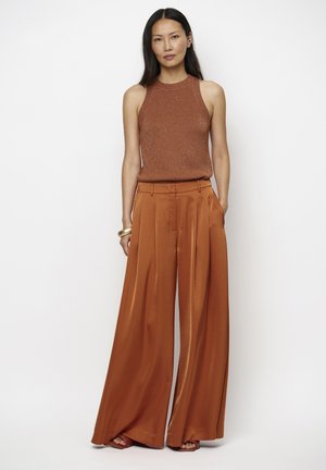 Vrouw draagt een roestkleurige mouwloze gebreide top en roestkleurige satijnen broek met wijde pijpen, staand met handen in de zakken tegen een effen witte achtergrond.