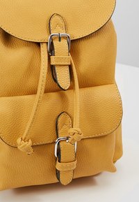 Žlutý texturovaný kožený batoh se dvěma stříbrnými přezkami a stahovací šňůrkou na bílé podložce.