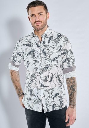 Weißes Hemd mit schwarzen botanischen Drucken, kurzen Ärmeln, Knopfkragen, Brusttasche und aufgerollten Manschetten. Der Stoff hat eine strukturierte Oberfläche.