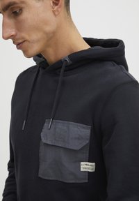 Tengerészkék kapucnis pulóver szürke zseb részletekkel, zsinórokkal és textil címkével. Simább textúra és laza kialakítás.