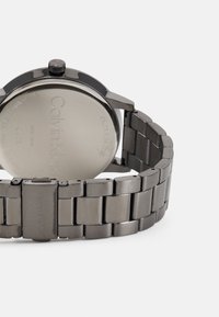Calvin Klein Uhr mit einem Armband aus rostfreiem Stahl in Dunkelgrau. Rundes Gehäuse mit einer glatten Oberfläche und graviertem Rückteil, das minimalistische Details aufweist.