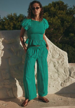 Nő, aki tengerzöld csíkos fodros ujjú felsőt és hozzáillő bő szárú nadrágot visel, szabadtéren áll egy fehér kőfal mellett, a háttérben fák láthatók.