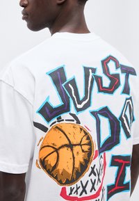 Άνδρας που φοράει λευκό μπλουζάκι με πολύχρωμο κείμενο "JUST DO IT" και μεγάλο πορτοκαλί γραφικό μπάσκετ στην πλάτη.