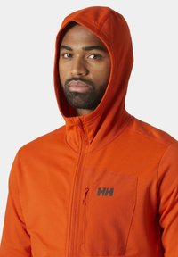 Pomarańczowa kurtka z kapturem o fakturowanej tkaninie i zamkiem błyskawicznym z przodu, z kieszenią na klatce piersiowej z wyszywanym czarnym logo.