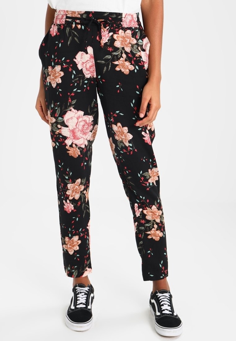 Person klädd i svarta byxor med blommönster i rosa och persika, i kombination med svarta och vita snörskor mot en vit bakgrund.