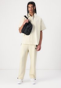 Ανοιχτό μπεζ κοντομάνικο πουκάμισο με κουμπί. Ταιριαστά μπεζ φόρμες με μαύρο λογότυπο Nike. Μαύρη τσάντα σώματος με λεπτομέρεια φερμουάρ.