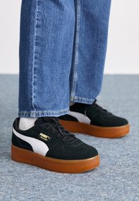 Baskets à plateforme en cuir de daim noir avec une bande blanche et un détail de marque doré, associées à un jean en denim bleu et des chaussettes blanches visibles.