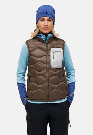 Kvinde iført en blå beanie, lyseblå og marineblå langærmet top, brun quiltet vest med hvid brystlomme og sorte bukser, hænderne i lommerne.