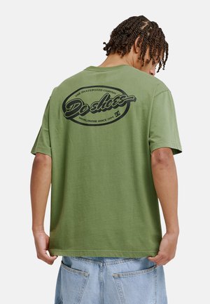 Oliwkowa bawełniana koszulka z krótkimi rękawami, z dużym czarnym okrągłym logo na plecach, na którym widnieje napis „DC Shoes” oraz „World Wide Since 1994.”
