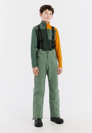 Groene waterdichte broek met verstelbare bretels en ritszakken, gecombineerd met een tweekleurig bovenstuk in groen en oranje. Heeft een hoge kraag.