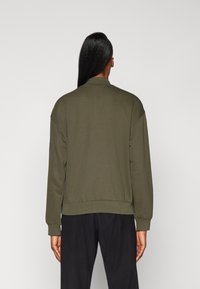 Olivgrüner Sweatshirt mit Kragen und gerippten Bündchen, lässiger Schnitt und glatte Textur, getragen über schwarzen Hosen.
