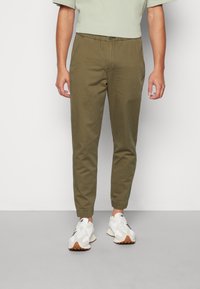 Pantalones jogger verde oliva con cintura elástica, puños en los tobillos y cierre frontal de botón, hechos de una tela ligera. Llevados con zapatillas blancas.