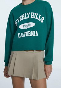 Felpa verde cropped con testo stampato bianco "BEVERLY HILLS CALIFORNIA EST. 1993." Abbinata a una gonna plissettata beige.