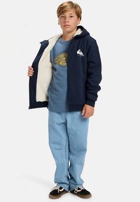 Navy blue zip-up hoodie with white fleece lining and logo, worn over a blue graphic t-shirt. Light blue jeans and black shoes complete the look.