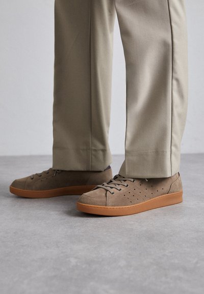 Brune ruskinds sneakers med perforerede detaljer og brune gummisåler, parret med lyse beige bukser, som står på en grå overflade.