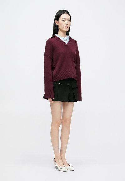 Bordeaux színű, oversized kapucnis pulóver, sötét szoknya oldalsó zsebekkel és fehér, hegyes orrú cipők. Simább textúrák és laza fit.