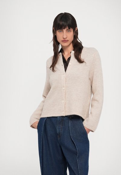 Sötét hajú nő bézs színű, gombos kardigánt visel fekete ing fölött, laza szabású sötétkék farmerral, kezét a zsebében tartva áll.