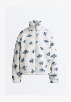 Bijela flis jakna s crno-sivim apstraktnim cvjetnim uzorcima, visokim ovratnikom i patentnim zatvaračem sprijeda.