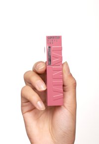 Maybelline Superstay Vinyl Ink, tubo rettangolare, rosa opaco, superficie testurizzata, logo frontale, tenuto in una mano.