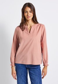 Helles rosa Langarmhemd mit V-Ausschnitt, gerafften Schulterdetails und elastischen Bündchen, kombiniert mit blauen Jeans.