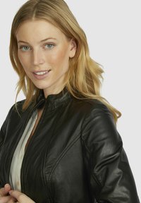 Black leather jacket with a fitted design. Features a high collar and subtle stitching details. Accompanied by a light-colored blouse underneath.