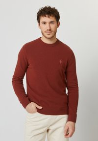 Maglione girocollo marrone rossastro realizzato in materiale morbido, con un piccolo logo ad albero sul petto e polsini e orlo a coste.